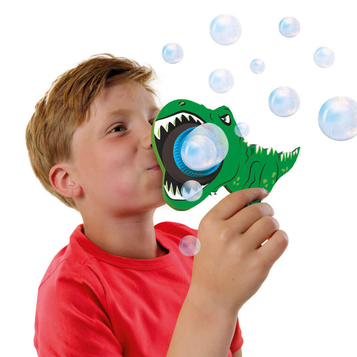 Child blowing bubbles through a green T-Rex shaped wand with clear bubbles floating against a white background.
