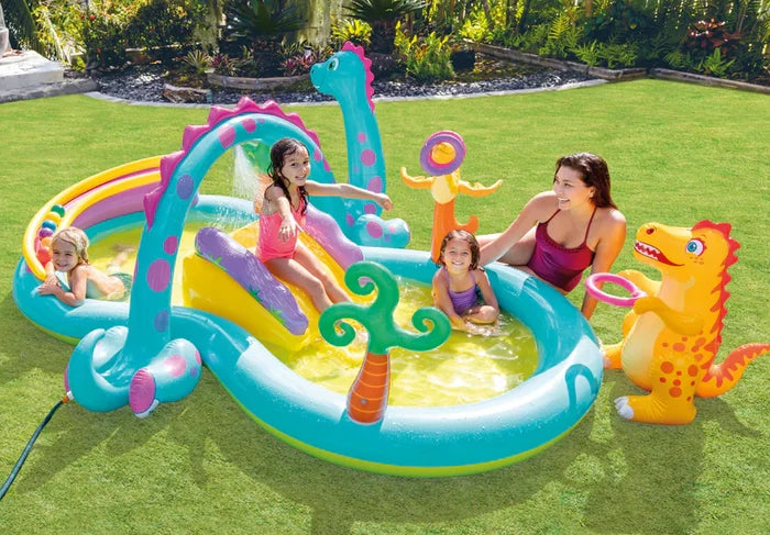 Teal and yellow inflatable dinosaur pool with slide, water sprayer, and hoop toss game on a green lawn.