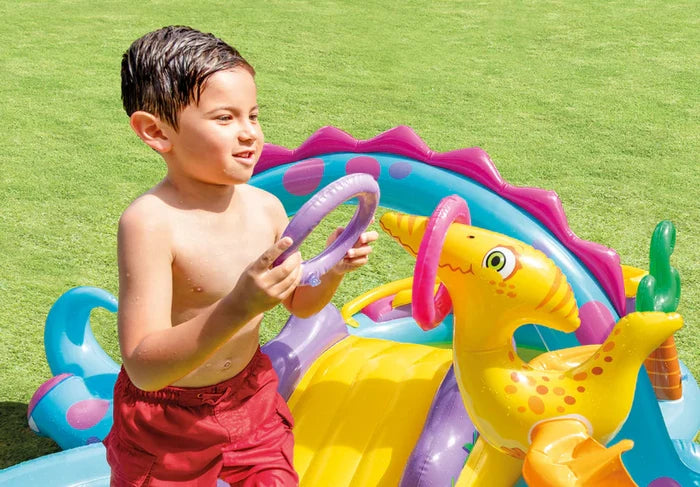 Boy playing with purple rings and a yellow dinosaur slide in a colorful inflatable paddling pool on a lawn.