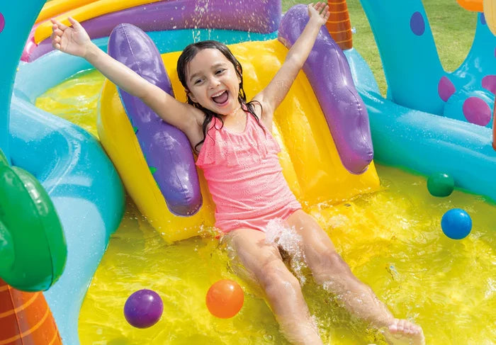 Girl in a pink swimsuit slides down a yellow inflatable slide into a blue pool with splashing water and colorful balls.