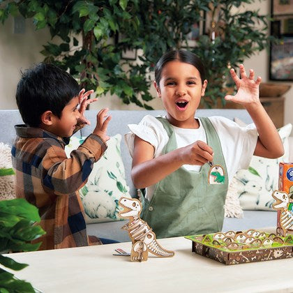 Two children roar while playing a dinosaur game with a 3D fossil model and a board with illustrated tokens.