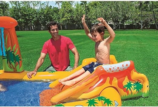 Boy sliding down a red and orange volcano-themed inflatable pool slide into a blue pool in a green yard.