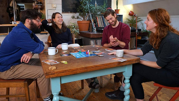 Four people laughing and playing a board game with colorful illustrated cards and game pieces on a wooden table.