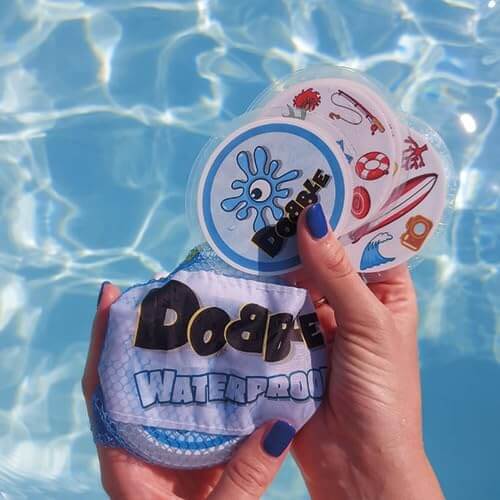 Hands hold circular waterproof Dobble cards with summer icons and a mesh storage bag over a blue swimming pool.
