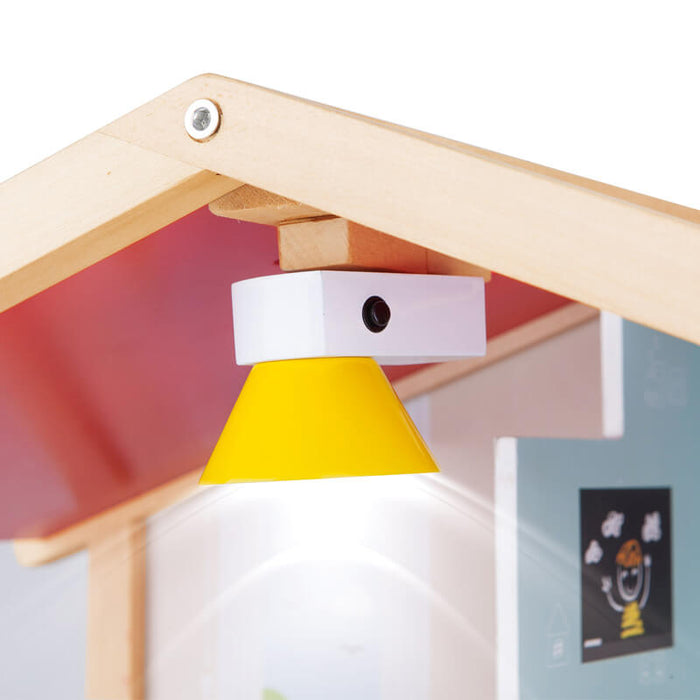 Glowing yellow conical lamp with a white base and black button, mounted to the wooden roof peak of a toy dollhouse.