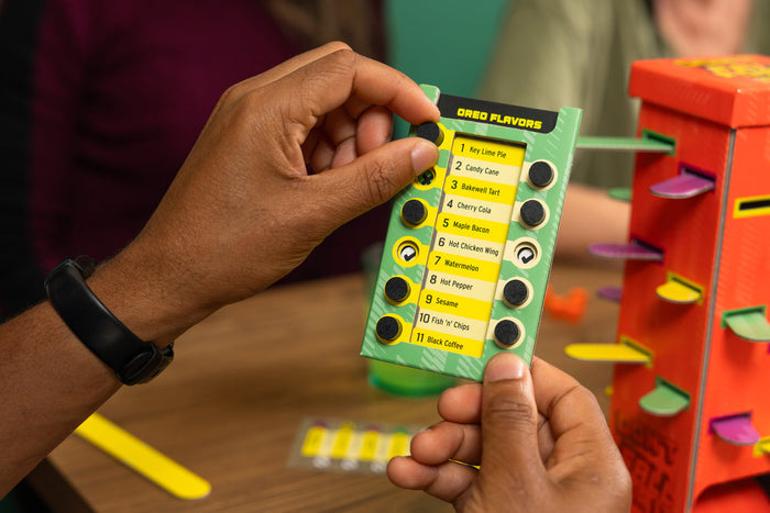Hands holding a green flavor card with black sliders next to a tall orange tower with colorful pegs.