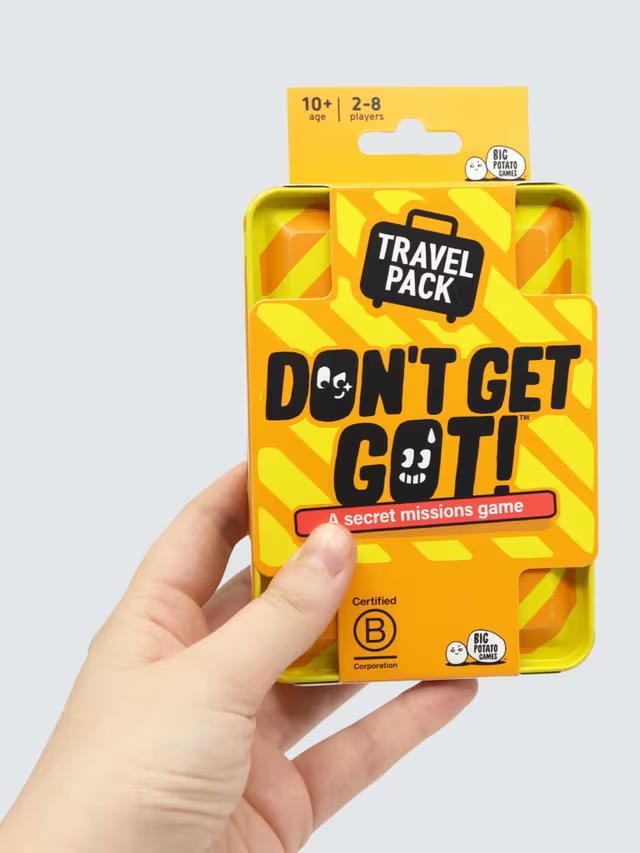 A hand holds an orange and yellow striped metal tin for the Don't Get Got Travel Pack secret missions game.