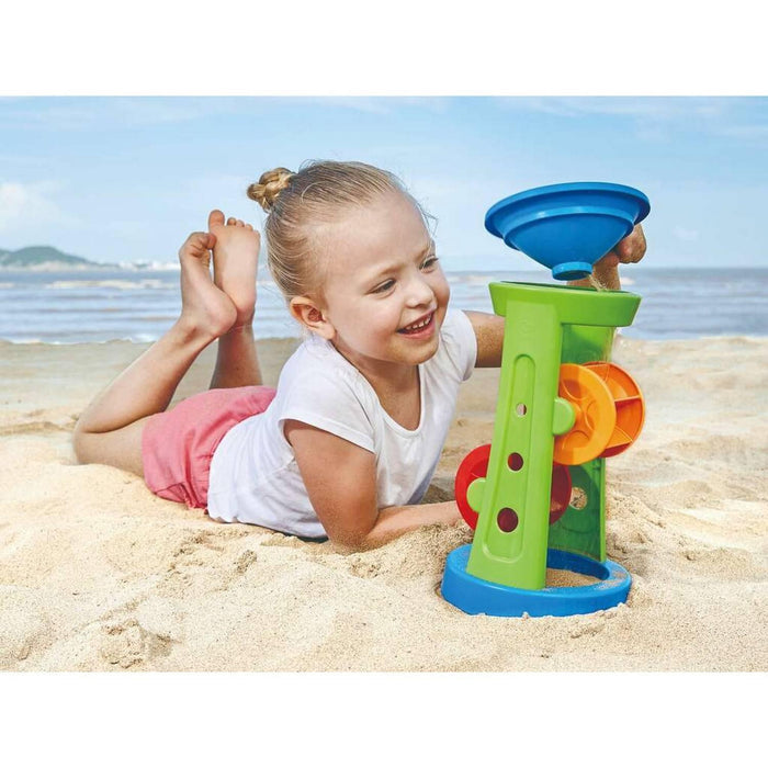 Child on a beach playing with a lime green toy tower, blue funnel, and spinning orange and red wheels.