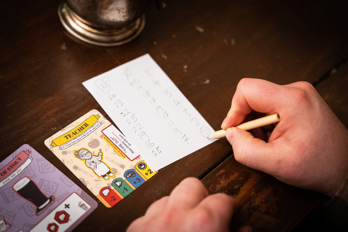 Hand marking scores on paper next to illustrated Teacher and Stout game cards on a dark wooden table.