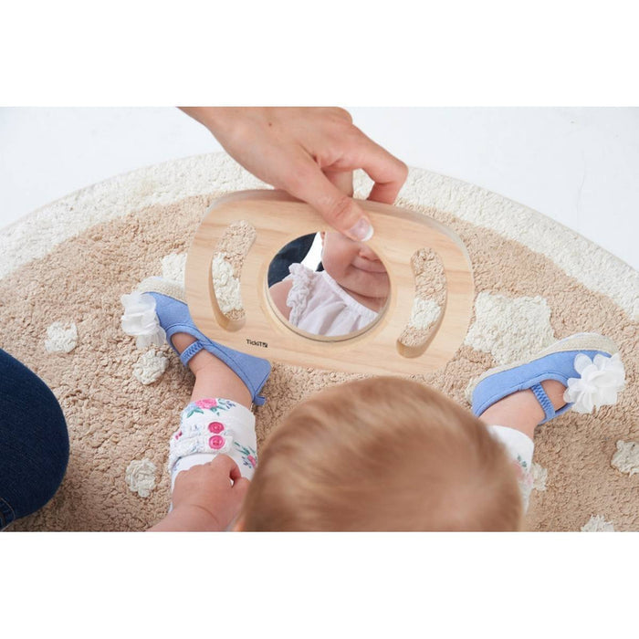 Oval wooden hand mirror with two easy-grip cutout handles, held by an adult in front of a seated baby.