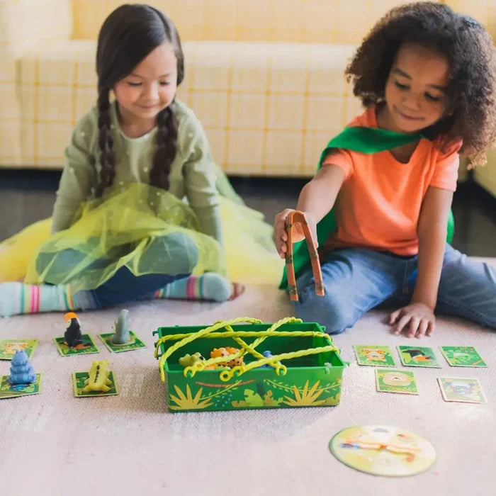 Two girls use orange tweezers to rescue toy animals from a green vine-covered box in a jungle board game.