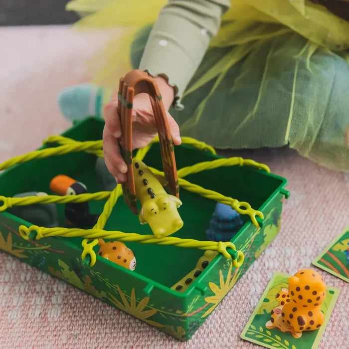 Child uses brown tongs to lift a toy crocodile from a green box with yellow vines and various jungle animal figures.