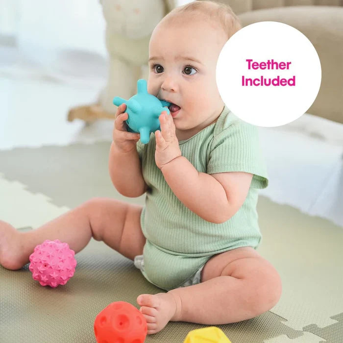 A baby bites a blue spiky sensory ball. A pink star ball and red dimpled ball sit on a gray play mat.