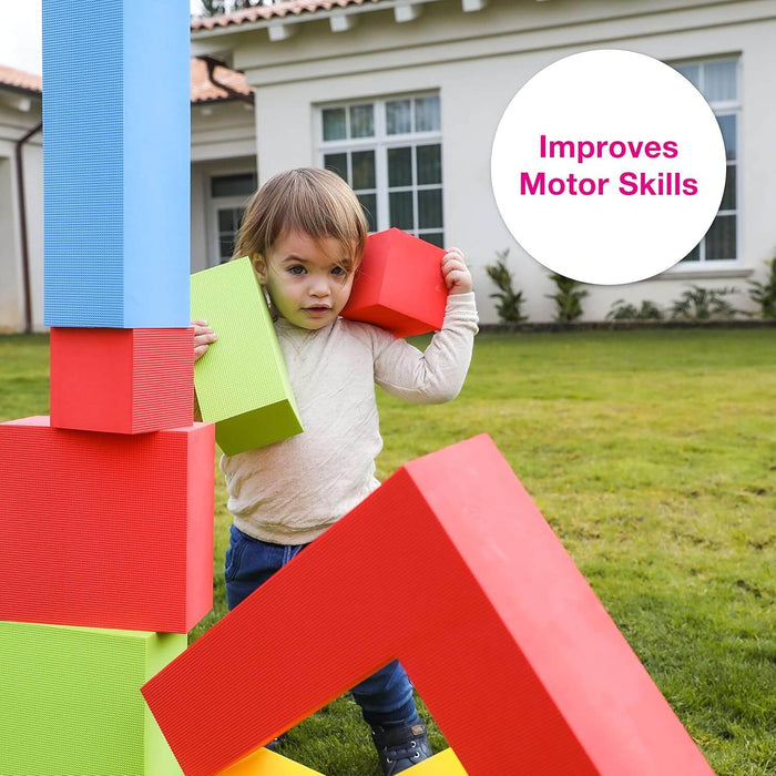Young child holding large red and green foam blocks beside a stack of colorful, textured blocks in a backyard.