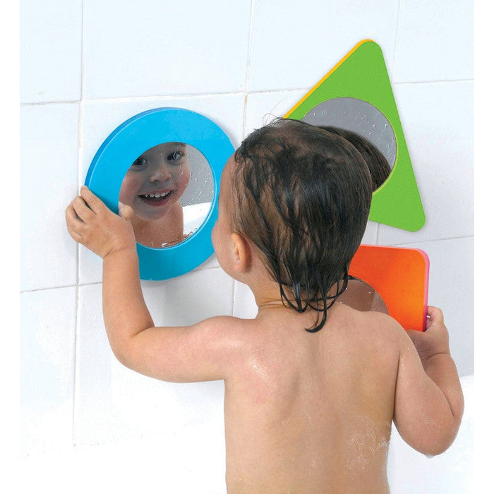 Toddler looks into a blue circular bath mirror next to a green triangle and orange square mirror on a tiled wall.