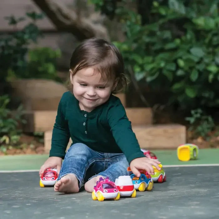 Toddler playing with bright, rounded toy cars featuring cartoon faces and yellow wheels on a dark outdoor surface.