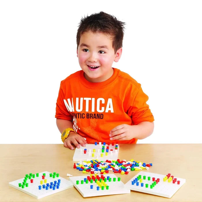 A young boy smiling while playing with colorful plastic pegs and white square pegboards on a wooden table.