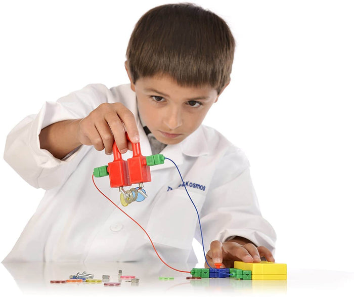 Boy in a lab coat uses a red electromagnet to pick up metal washers, connected by wires to a yellow battery box.