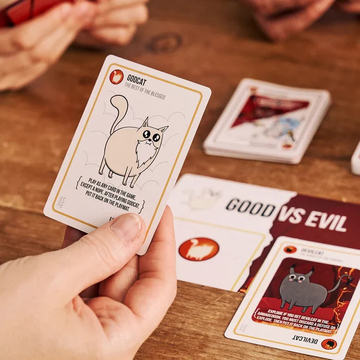 Hand holding a white Godcat card with gold trim over a Good vs Evil playmat and red Devilcat card.