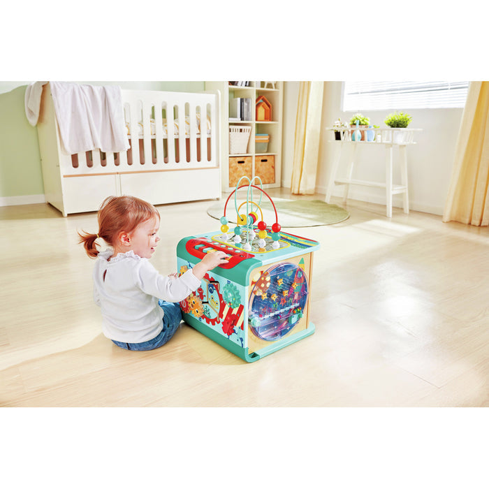 Toddler playing with a colorful multi-activity wooden cube featuring a bead maze, gears, and lights.