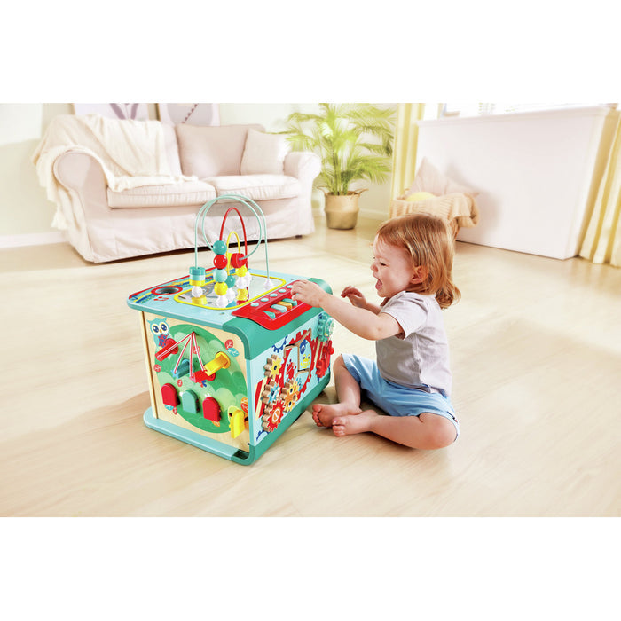 Teal wooden activity cube with bead maze and spinning gears, shown with a toddler playing in a bright room.