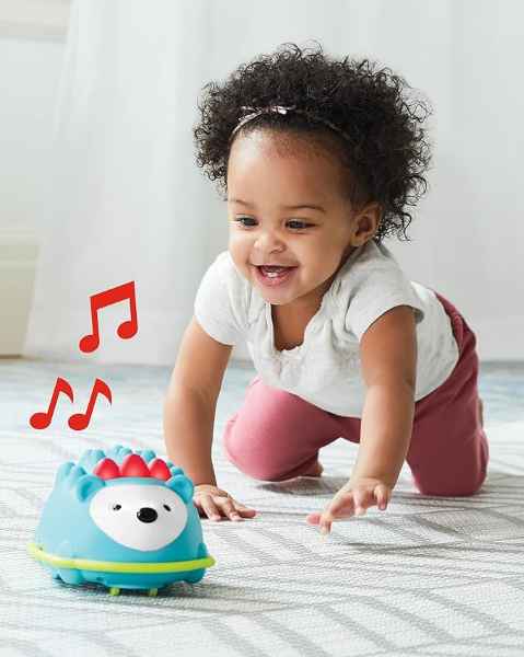 Baby crawling on a grey rug towards a blue hedgehog toy with red light-up spikes and musical note graphics.