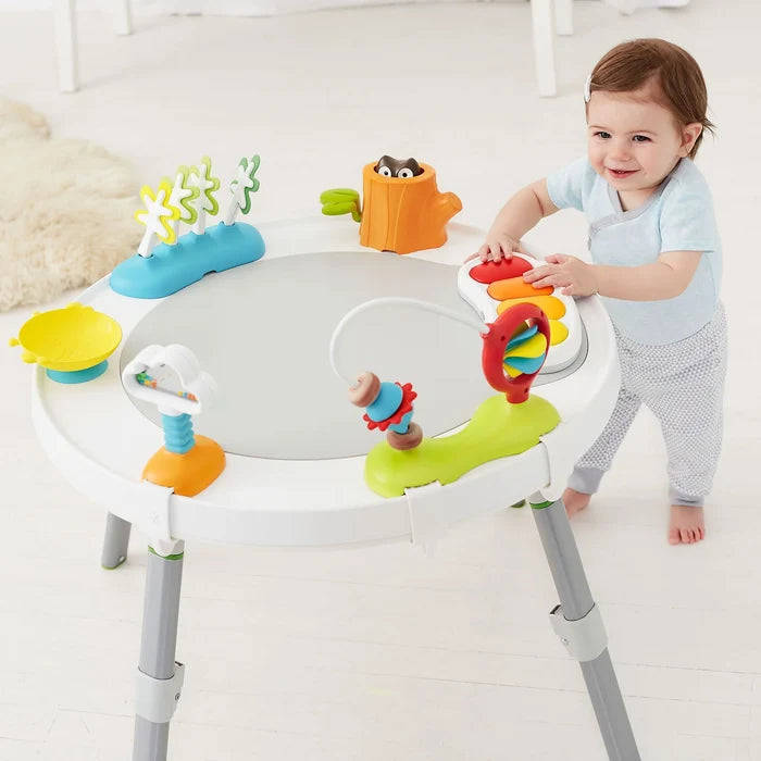 Baby stands at a round white activity table with colorful toys including a light-up keyboard and owl tree stump.