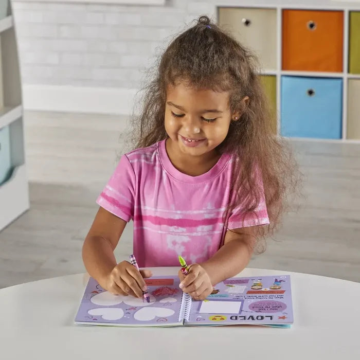 Smiling young girl coloring in a colorful spiral-bound activity journal with crayons at a white table.