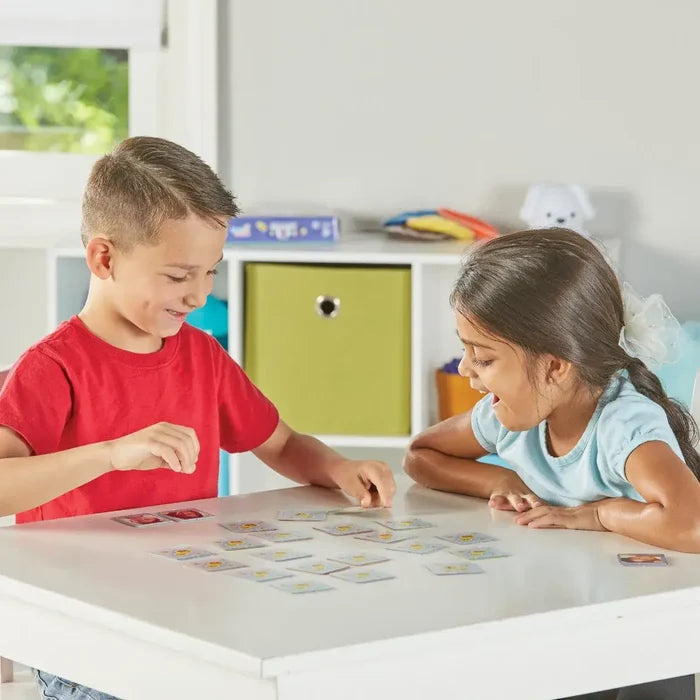 Two children play a memory game with square cards featuring various facial expressions on a white table.