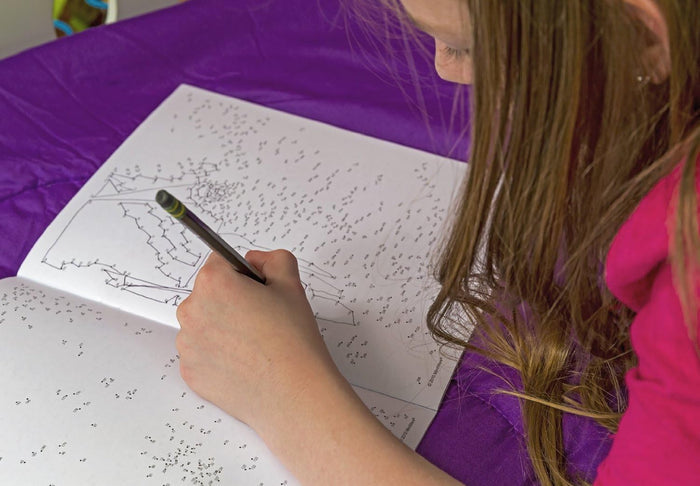 Close-up of a child drawing in a complex dot-to-dot book with a gray pencil on a purple surface.