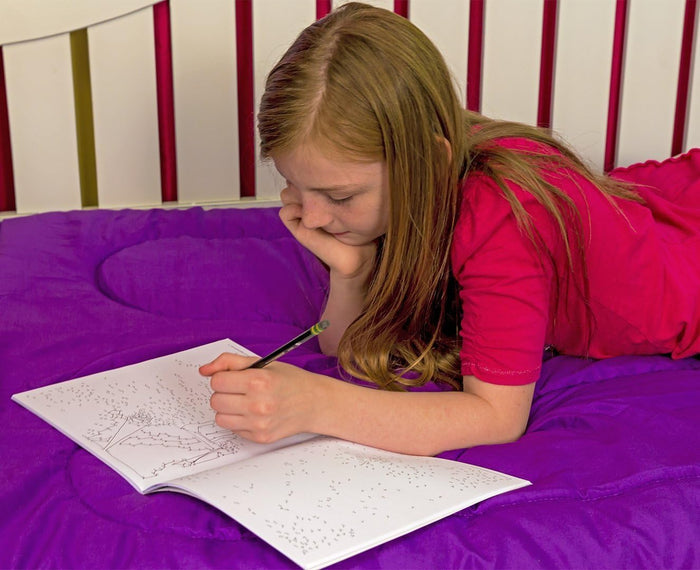 Young girl lying on a purple comforter, connecting complex dot-to-dot patterns in an open book with a pencil.