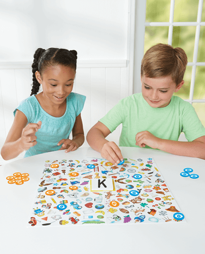 Boy and girl play a board game with colorful illustrations, blue and orange eye tokens, and a letter K card.