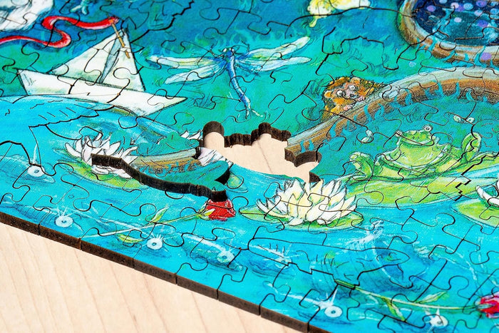 Close-up of a wooden jigsaw puzzle with whimsical piece shapes and a missing piece, featuring a colorful pond scene.