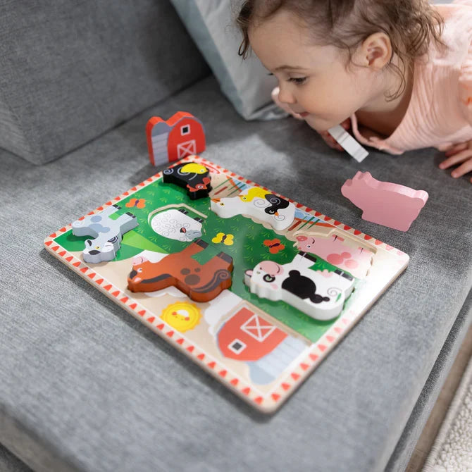 Toddler looks at a colorful wooden farm puzzle with chunky animal pieces like a cow, horse, and pig on a grey sofa.