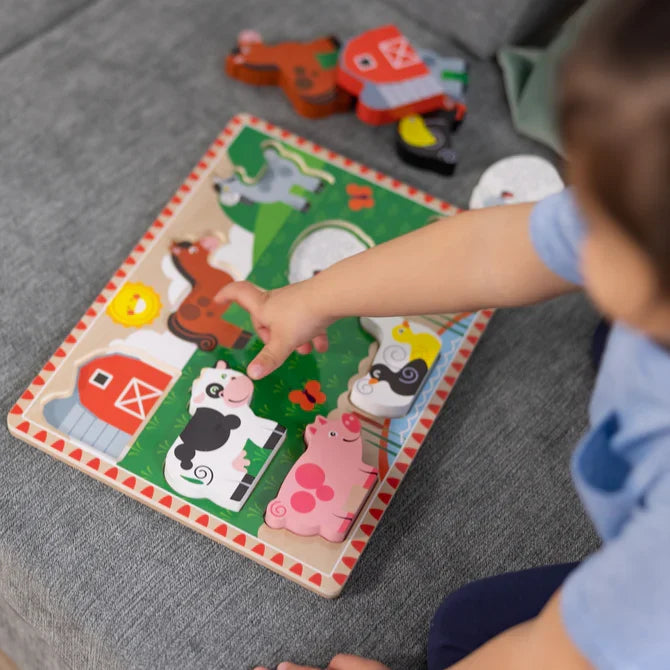 Child points to a chunky wooden cow piece on a farm-themed puzzle featuring a pink pig, brown horse, and red barn.