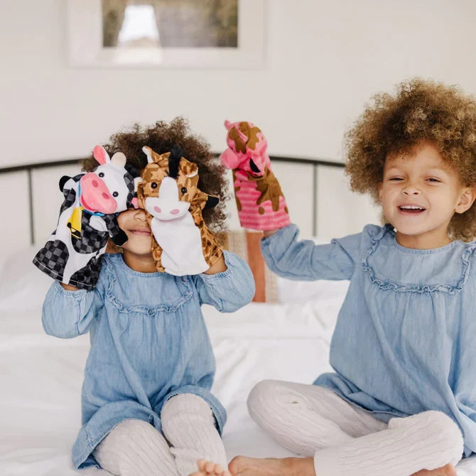 Two girls playing with plush cow, horse, and pig hand puppets on a white bed.