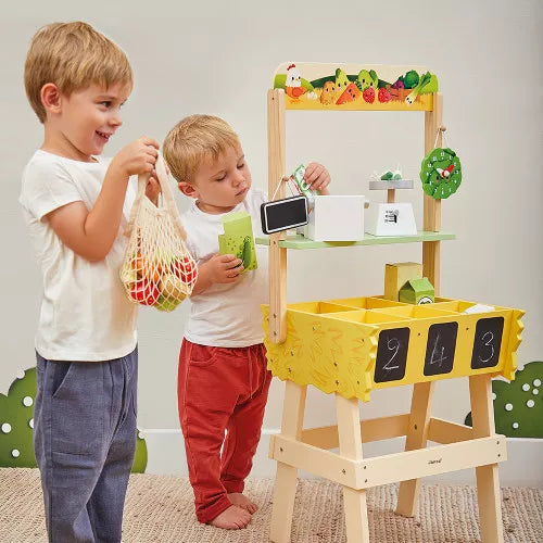 Two boys playing with a wooden farm market stall featuring colorful fruit graphics, a toy scale, and chalkboards.