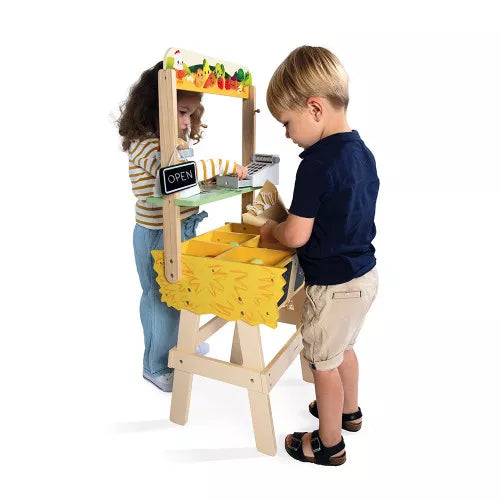 Two children play with a wooden toy market stall featuring yellow produce bins, a toy cash register, and an open sign.