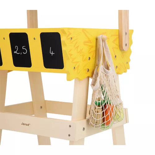 Wooden toy market stall with a yellow bin, chalkboards, and a mesh bag of wooden vegetables hanging on the side.
