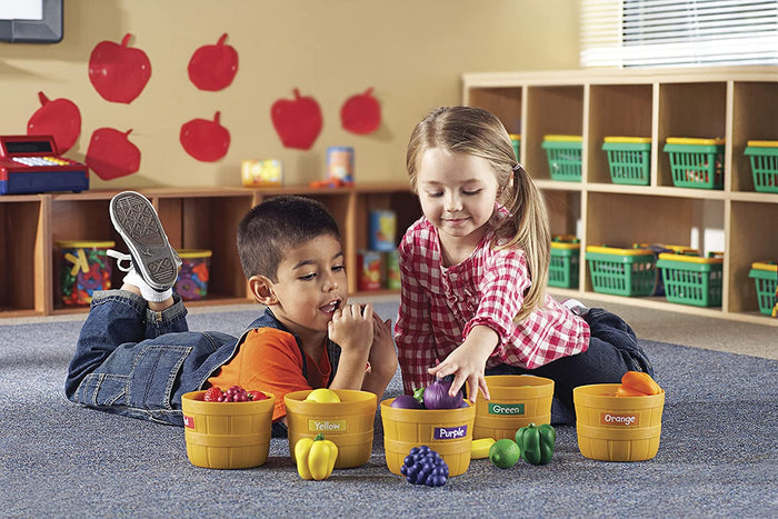 Two kids sorting colorful toy produce into five yellow baskets labeled by color on a blue classroom rug.