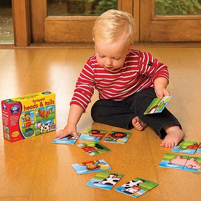 Toddler in a red striped shirt matches farm animal cards on a wooden floor next to the yellow Orchard Toys box.