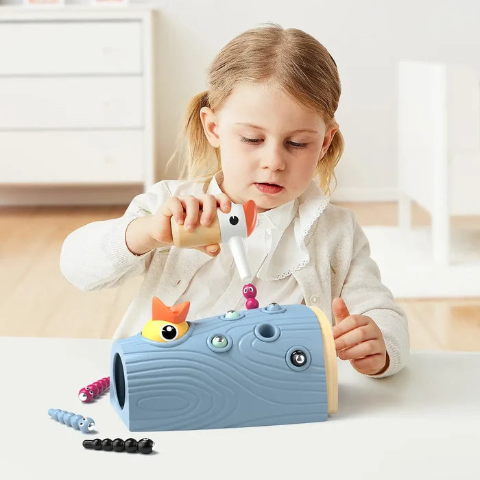 Child uses a bird tool to pull colorful magnetic worms from a blue textured log toy to feed a yellow baby bird.