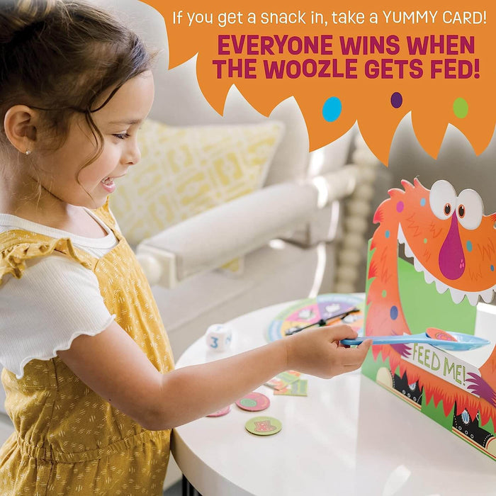 A young girl uses a blue spoon to feed snacks into an orange monster's mouth. Game tokens and a die are on the table.