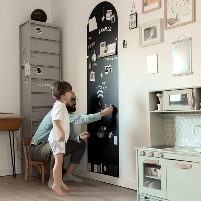 Tall arched black magnetic wallpaper strip with photos and chalk, being used by a man and young boy.