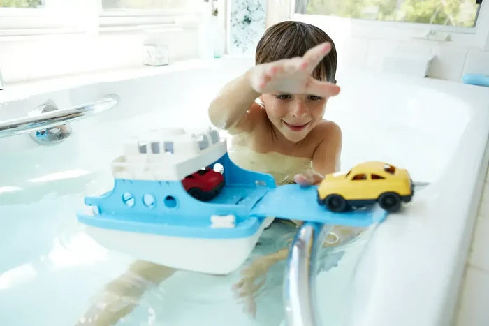 A child in a bathtub plays with a blue and white toy ferry boat carrying a red car and a yellow car on its ramp.