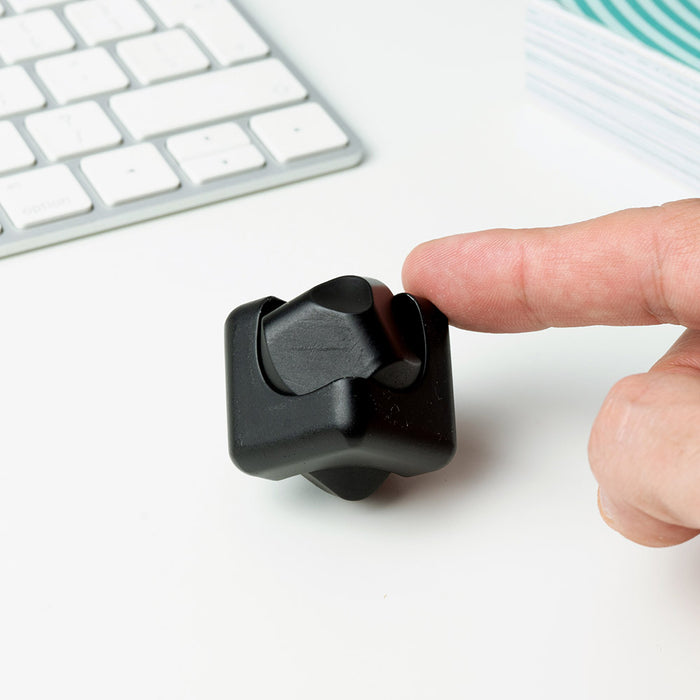 Matte black metal spinning cube toy on a white desk being touched by a finger, near a computer keyboard.