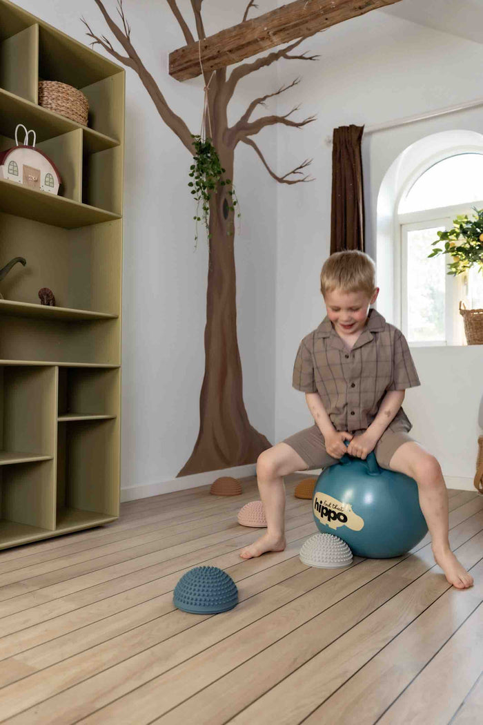 Child sitting on a dark cyan hopper ball with a handle and logo on a light wood floor in a neutral playroom.