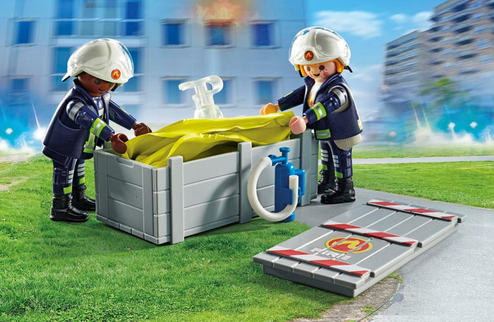 Two toy firefighters setting up a large yellow air pillow in a grey container with a blue pump on green grass.
