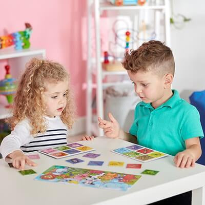 Two children play a colorful animal matching game with square tiles and boards on a white table.