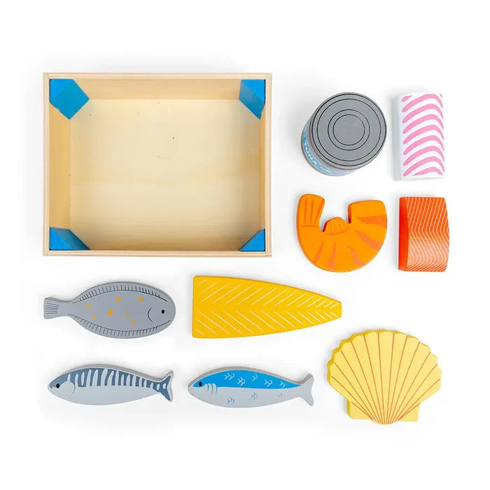 Wooden play seafood set with various painted fish, fillets, a shrimp, shell, and tuna can next to a wooden crate.